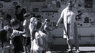 Pfarrer Alfons Dirscherl inmitten der Kinder mit Lehrerin Elfriede Bogner (links) bei der Einweihung des Wandfries an der Mauer der Grundschule, das im Schuljahr 2018/19 zusammen mit den beiden K&uuml;nstlern Georg Fuchssteiner und Peter Herr (Bogen) geschaffen wurde.