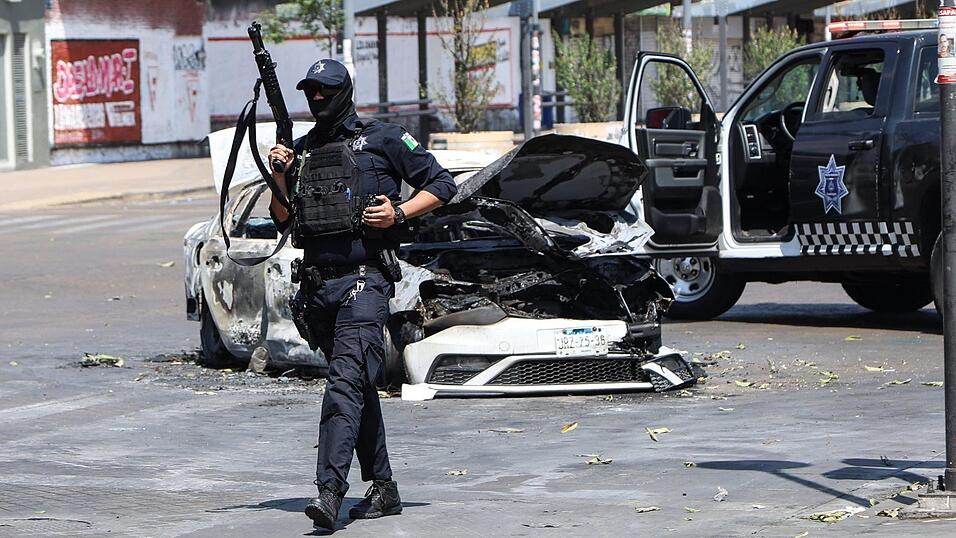Ein Polizist steht in Guadalajara, der Hauptstadt des mexikanischen Bundesstaates Jalisco, neben einem verbrannten Auto auf der Stra&szlig;e Wache. Am vergangenen Wochenende gab es wegen der T&ouml;tung des Drogenbosses &bdquo;El Mencho&ldquo; heftige Unruhen mit vielen Toten.