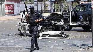 Ein Polizist steht in Guadalajara, der Hauptstadt des mexikanischen Bundesstaates Jalisco, neben einem verbrannten Auto auf der Stra&szlig;e Wache. Am vergangenen Wochenende gab es wegen der T&ouml;tung des Drogenbosses &bdquo;El Mencho&ldquo; heftige Unruhen mit vielen Toten.