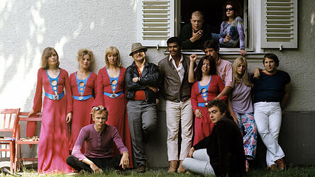 Bürgerschreck und Kult-Regisseur Rainer Werner Fassbinder (stehend, 4.v.l.) 1970 mit Mitgliedern des Münchner Anti-Theater-Ensembles, darunter (v.l. stehend): Margit Carstensen, Hanna Schygulla, Kerstin Dobbertin, R. W. Fassbinder, Günter Kaufmann, Harry Bär, davor (im roten Kleid) Ursula Strätz und vor ihr sitzend Kurt Raab; rechts Fassbinders enger Freund Peer Raben, der die Musik zu 26 seiner Filme machte. Im Fenster Rudolf Waldemar Brem (l.) und eine unbekannte Frau.