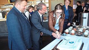 Unter den Augen des zweiten B&uuml;rgermeisters (links) und von Hauptgesch&auml;ftsf&uuml;hrerin Ute Mowitz-Rudolph (rechts) schneiden Verbraucherberaterin Nicole Br&auml;u und Minister Glauber die Geburtstagstorte an.