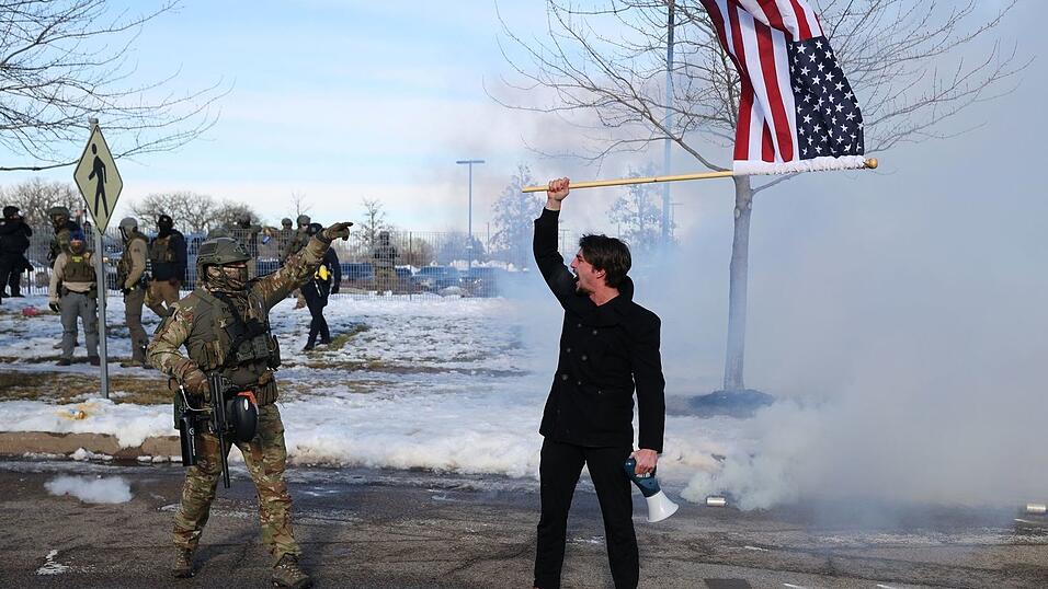 Ein Protestierender mit einer amerikanischen Flagge steht einem ICE-Beamten gegen&uuml;ber.