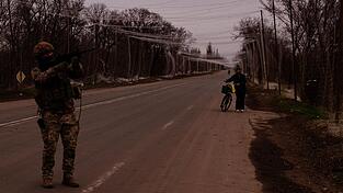 &Uuml;ber der Stra&szlig;e h&auml;ngt ein Netz aus d&uuml;nnen F&auml;den, das Sprengstoff-Drohnen abhalten soll. Ruslan posiert mit seinem Gewehr, eine Frau wei&szlig; nicht so recht, wo sie mit ihrem Rad hinsoll. Nur noch wenige Zivilisten sind in den oft in gro&szlig;en Teilen zerst&ouml;rten D&ouml;rfern geblieben.