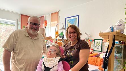 Henry und Yvonne Dinger mit ihrer Tochter Katharina in ihrem Wohnzimmer. Der Esstisch neben dem Tagesbett ist gleichzeitig der Schreibtisch, das erleichtert den Eltern die Pflege ihrer Tochter, wenn sie arbeiten. Ein Team der Spezialisierten Ambulanten Palliativversorgung der LA-Regio Klinken begleitet die Familie in ihrer Lebenssituation.