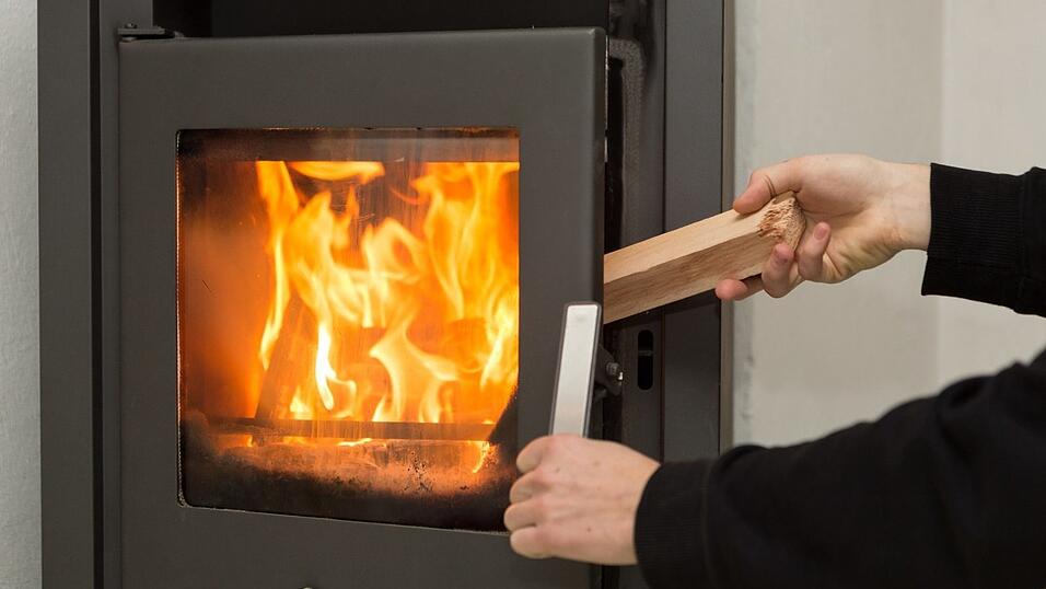 Ein Mann legt in seiner Wohnung ein Stück Holz in einen Kaminofen, in dem ein Feuer brennt. Ein Mann legt in seiner Wohnung ein Stück Holz in einen Kaminofen, in dem ein Feuer brennt.