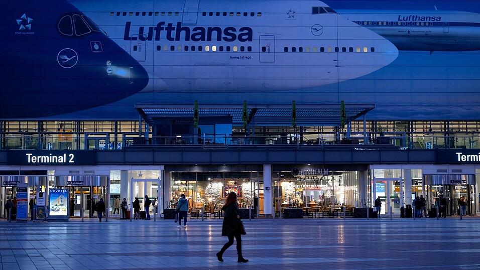 Passagiere gehen am M&uuml;nchner Flughafen vor dem Terminal 2 an einem Plakat mit Flugzeugen der Lufthansa entlang.