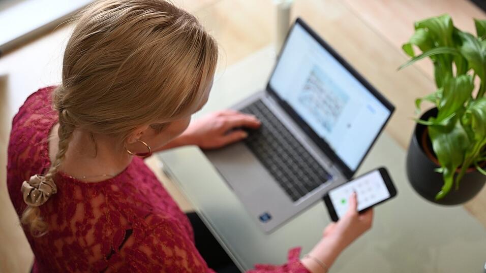 Eine Frau sitzt zuhause an einem Glastisch und tippt auf einem Laptop, in der rechten Hand hält sie ein Smartphone. Eine Frau sitzt zuhause an einem Glastisch und tippt auf einem Laptop, in der rechten Hand hält sie ein Smartphone.