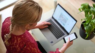 Eine Frau sitzt zuhause an einem Glastisch und tippt auf einem Laptop, in der rechten Hand hält sie ein Smartphone.