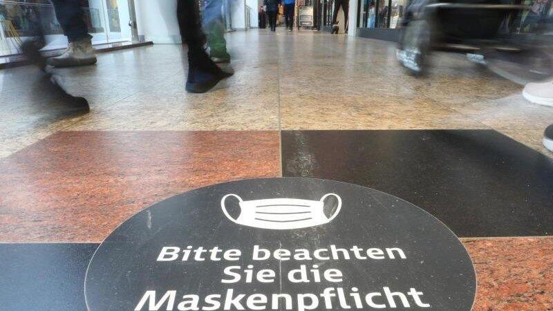 Passanten laufen durch ein Einkaufszentrum auf dessen Fußboden ein Hinweisschild zur Maskenpflicht aufgeklebt ist.
