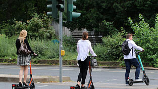 Ein Mann und zwei junge Frauen überqueren mit ihren E-Scootern an einer Ampel die Straße. Um einen solchen elektrischen Tretroller zu fahren, muss man mindestens 14 Jahre alt und das Gerät versichert sein. - Verkehrsexperten raten außerdem dazu, einen Helm zu tragen.