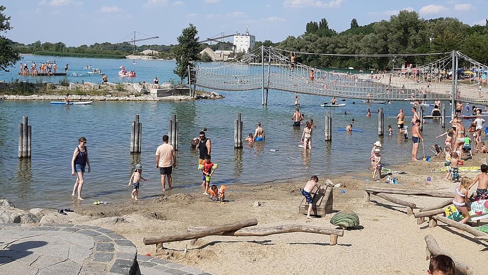 Der Sandstrand ist vor allem bei kleinen Kindern beliebt. Bis zu den Holzphählen geht der Nichtschwimmerbereich. Im Hintergrund eine der Naturinseln, die durch eine seit 2021 gesperrte Hängebrücke mit dem Ufer verbunden ist. Zur Holzinsel weiter draußen muss man schwimmen. Am Horizont sieht man das Kiesabbauwerk, das dem See seinen Namen gibt. Der Sandstrand ist vor allem bei kleinen Kindern beliebt. Bis zu den Holzphählen geht der Nichtschwimmerbereich. Im Hintergrund eine der Naturinseln, die durch eine seit 2021 gesperrte Hängebrücke mit dem Ufer verbunden ist. Zur Holzinsel weiter draußen muss man schwimmen. Am Horizont sieht man das Kiesabbauwerk, das dem See seinen Namen gibt.