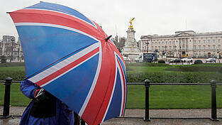 Ein Kameramann sch&uuml;tzt sich mit einem Regenschirm in den Farben der Nationalflagge des Vereinigten K&ouml;nigreichs, w&auml;hrend er den Buckingham-Palast filmt. Der Palast ringt nach dem Interview darum, die Kontrolle &uuml;ber eine eskalationsbereite Monarchiekrise zu behalten.