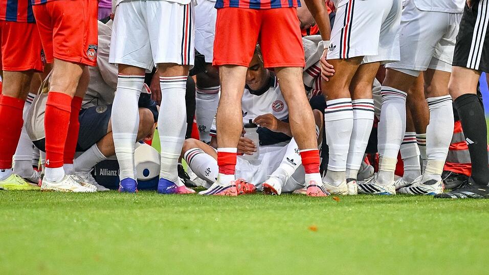 Jamal Musiala sitzt, abgeschirmt von M&uuml;nchner Teamkollegen und Heidenheimer Fu&szlig;ballern, nach seinem Zusammenbruch auf dem Rasen.