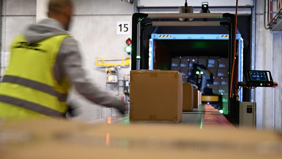 Ein DHL-Mitarbeiter nimmt Pakete in Emfpang - im Hintergrund ist ein Roboter zu sehen, der die Pakete aus einem Container nimmt und sie auf ein Förderband legt. (Archivbild) Ein DHL-Mitarbeiter nimmt Pakete in Emfpang - im Hintergrund ist ein Roboter zu sehen, der die Pakete aus einem Container nimmt und sie auf ein Förderband legt. (Archivbild)