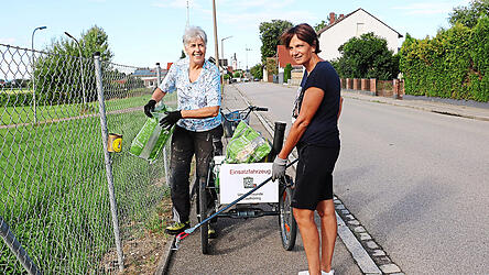 Brigitte Neuhäusler (links) hängt an einen Zaun eine Mülltüte, Brigitte Karl sammelt mit einer Greifzange Plastikabfälle vom Boden. Alle zwei Wochen streifen die beiden mit ihren Rädern durch Geiselhöring, um für eine sauberere Umwelt zu sorgen.