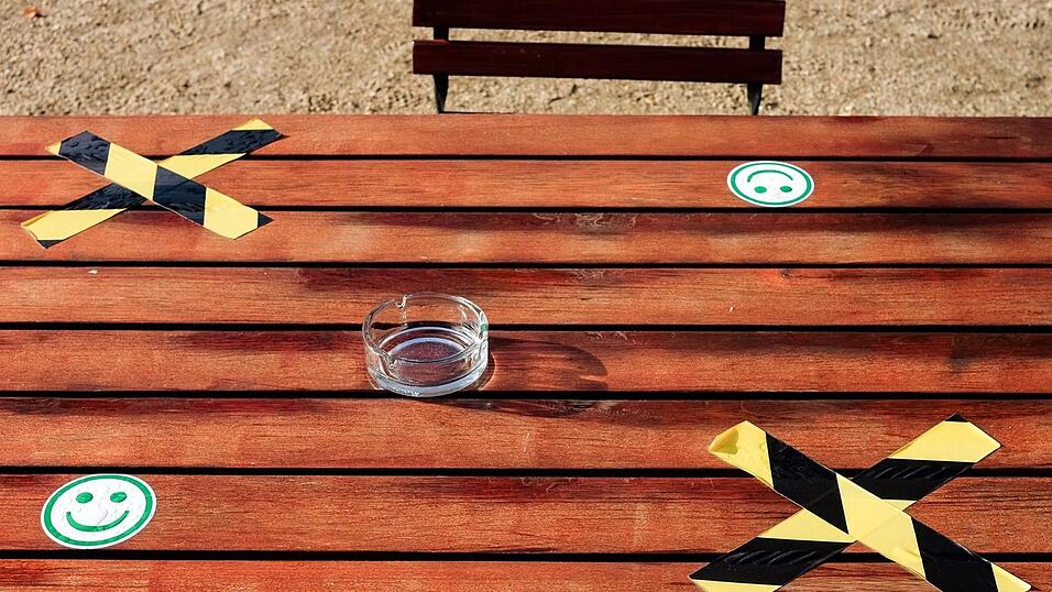 Auf einem Tisch eines Freiluftcafes steht zwischen aufgeklebten Kreuzen und Smileys für den Corona-Abstand ein leerer Aschenbecher. Auf einem Tisch eines Freiluftcafes steht zwischen aufgeklebten Kreuzen und Smileys für den Corona-Abstand ein leerer Aschenbecher.
