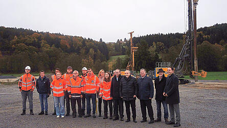 Gruppenbild mit Baumaschinen: Projektverantwortliche, Projektausführende, Regierungsmitarbeiter, Straßenbauamtsleiter und Mandatsträger auf dem Gelände der künftigen Ortsumgehung Auerbach.