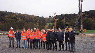 Gruppenbild mit Baumaschinen: Projektverantwortliche, Projektausführende, Regierungsmitarbeiter, Straßenbauamtsleiter und Mandatsträger auf dem Gelände der künftigen Ortsumgehung Auerbach.