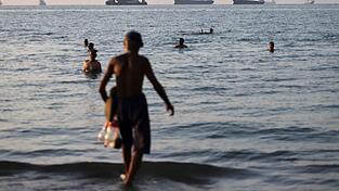 Anwohner baden im Wasser, während Fracht- und Handelsschiffe in der Straße von Hormus vor Anker liegen. Anwohner baden im Wasser, während Fracht- und Handelsschiffe in der Straße von Hormus vor Anker liegen.