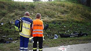 Ein Seelsorger und ein Mitglied der Feuerwehr an einer Unfallstelle auf der Autobahn 9 vor einer Böschung, die mit Trümmerteilen übersäht ist.