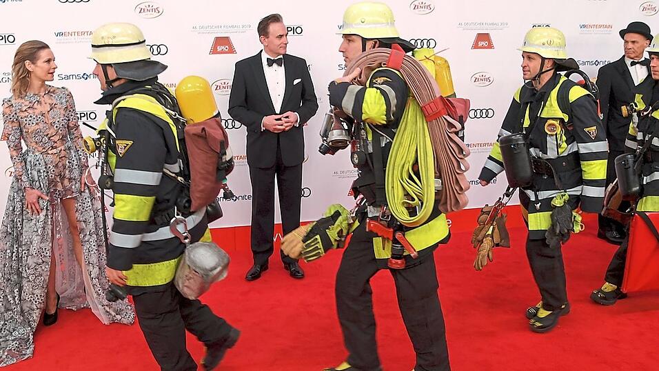 Nanu, sind das Stuntmen? Unter den &uuml;berraschten Blicken von Lisa Maria Martinek, ihrem Mann Giulio Ricciarelli und Heiner Lauterbach (r., mit Hut) laufen Feuerwehrleute beim Filmball &uuml;ber den roten Teppich vorm Bayerischen Hof. Die Feuerwehr ist wegen eines Fehlalarms gekommen.