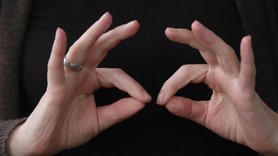 Eine Gebärdendolmetscherin zeigt mit ihren Fingern das Wort 'regelmäßig' in Gebärdensprache. Kosten für Gebärdendolmetscher gehören zu den Ausgaben, die ein regelmäßiges Gehörlosengeld abdecken soll.