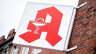 Ein Schild mit dem Apotheken-Logo hängt an einer Hausfassade.