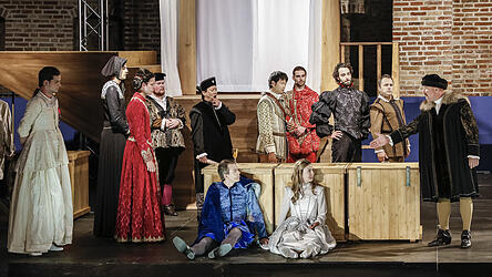 Will und Viola (Paul Behrens und Larissa Sophia Farr, sitzend) mit Schauspieler, Geldgeber und Theaterleitern auf der Bühne, von wo der Oberzensor sie vertreiben will.